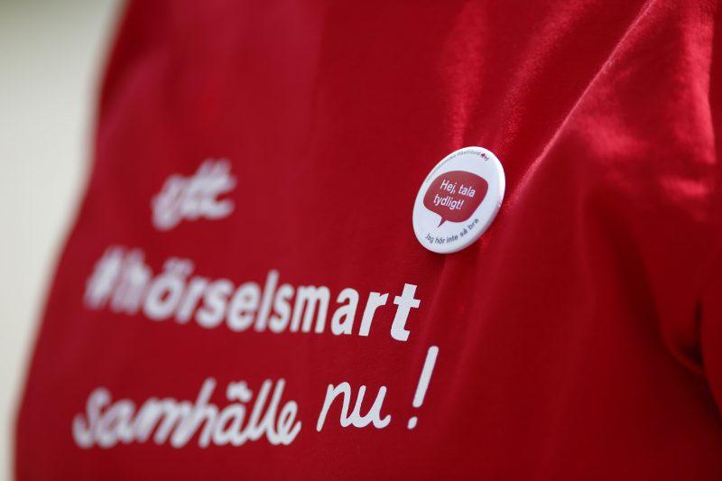 Närbild på t-shirt med knapp. Budskap: "Ett hörselsmart samhälle nu!"