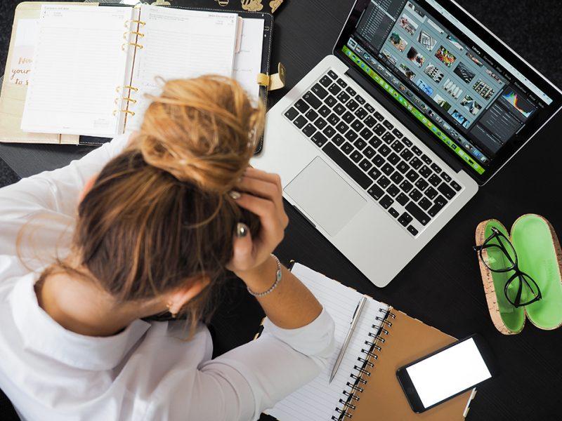 Kvinna framför laptop med huvudet i händerna, sedd uppifrån, bakifrån.