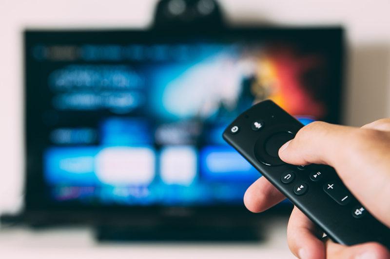 Hand håller fjärrkontroll framför suddig tv-skärm.