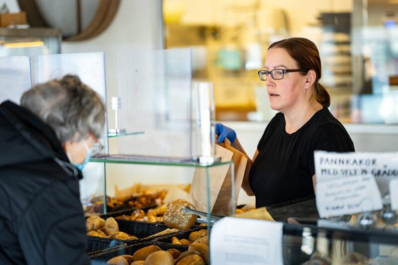 Kvinna med mörkt hår och glasögon bakom disk tittar koncentrerat på äldre kvinna med munskydd.