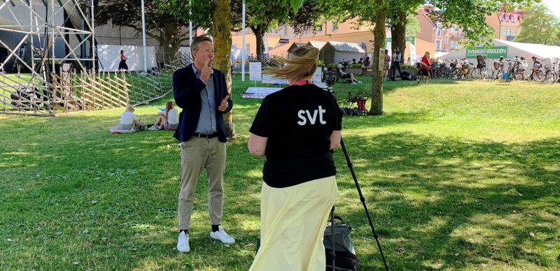 Kvinna i gul kjol och svart tröja med bokstäverna SVT står med filmkamera framför man i kavaj och som tecknar. Omgivning: Grön gräsmatta i park.