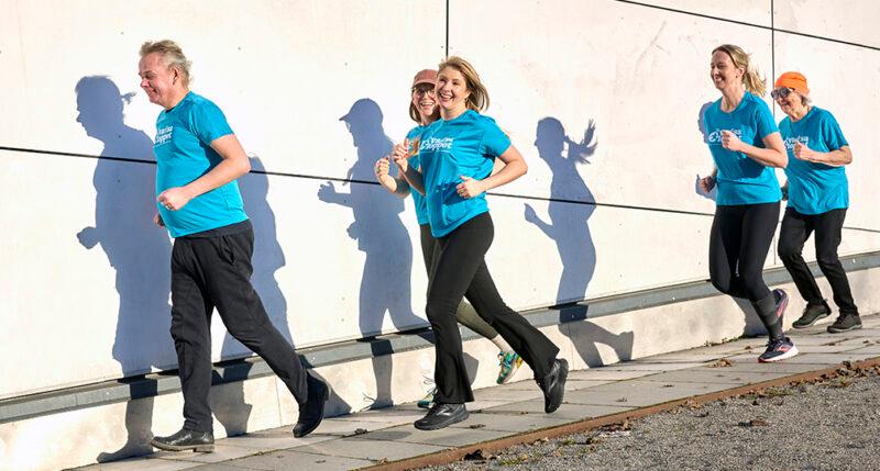 Fem personer i turkosblå tröjor joggar intill en vit vägg.