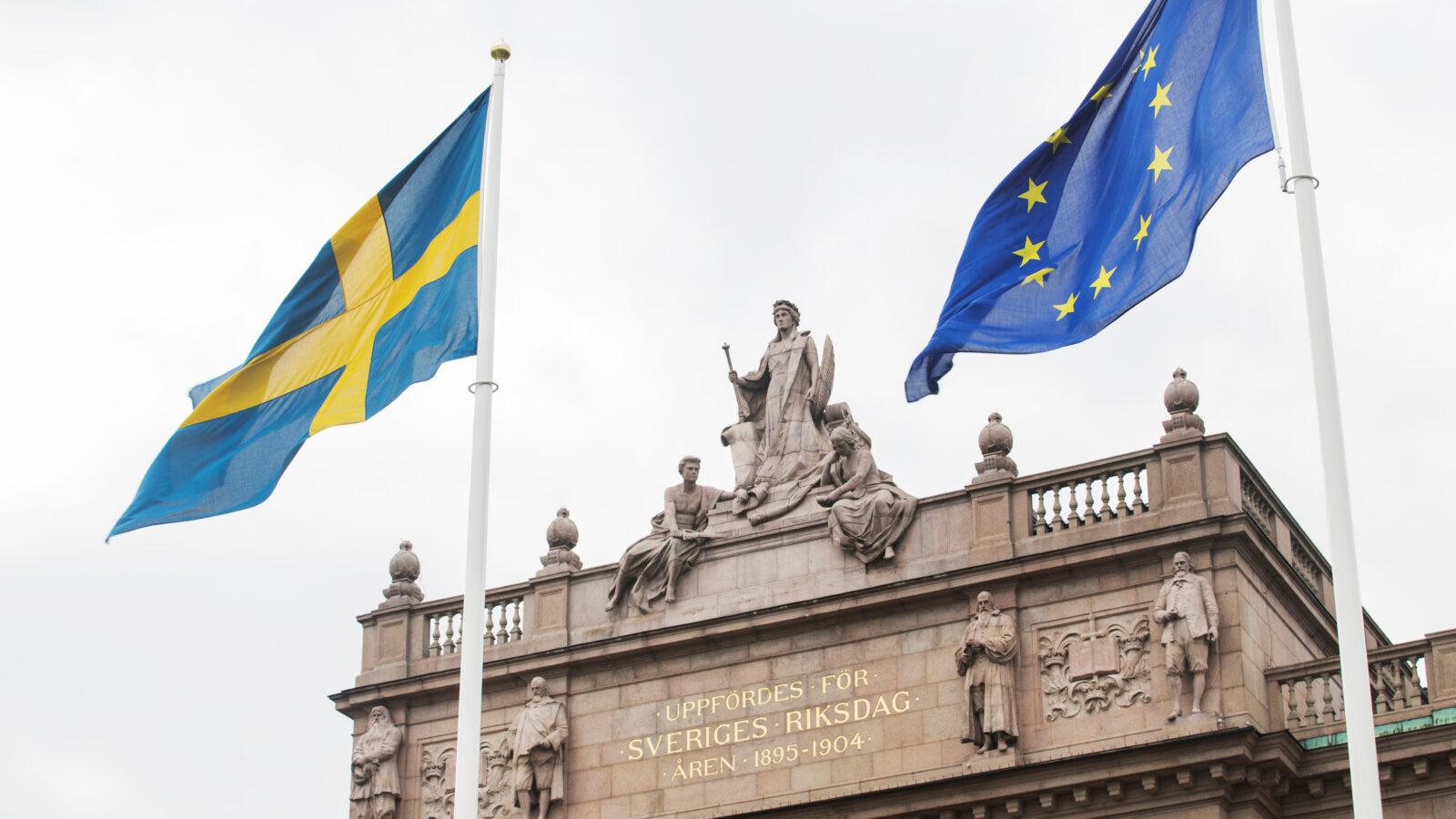 Del av riksdagshuset, med en svensk flagga och en EU-flagga framför.