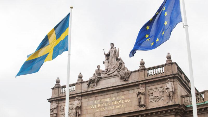 Del av riksdagshuset, med en svensk flagga och en EU-flagga framför.