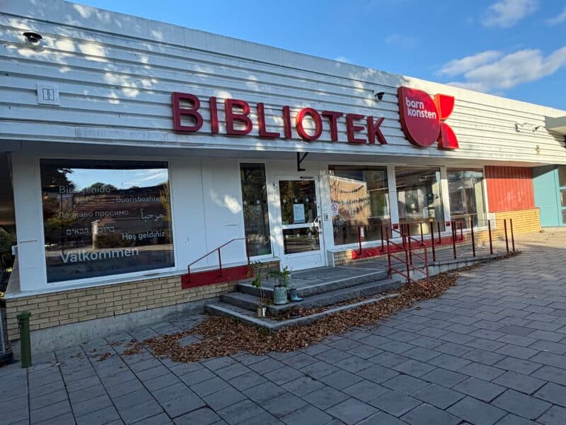 Bild på utsidan av Vårby bibliotek. Man ser entrén med ramp för rullstol eller barnvagn. I fönstret finns texter på olika språk som säger "Välkommen!"
