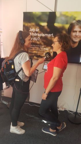 Radioreporter med mikrofon och sändningsutrustning på ryggen intervjuar kvinna i röd HRF-tröja.
