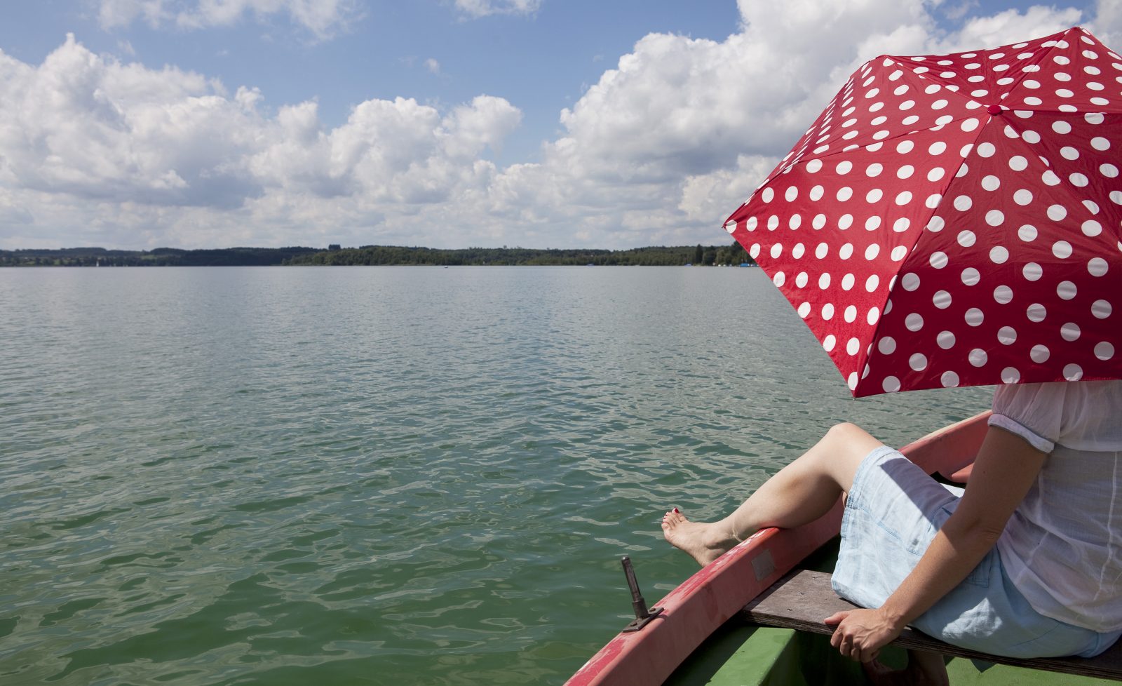 Kvinna sitter vid stilla sommarsjö, i roddbåt med rött parasoll.