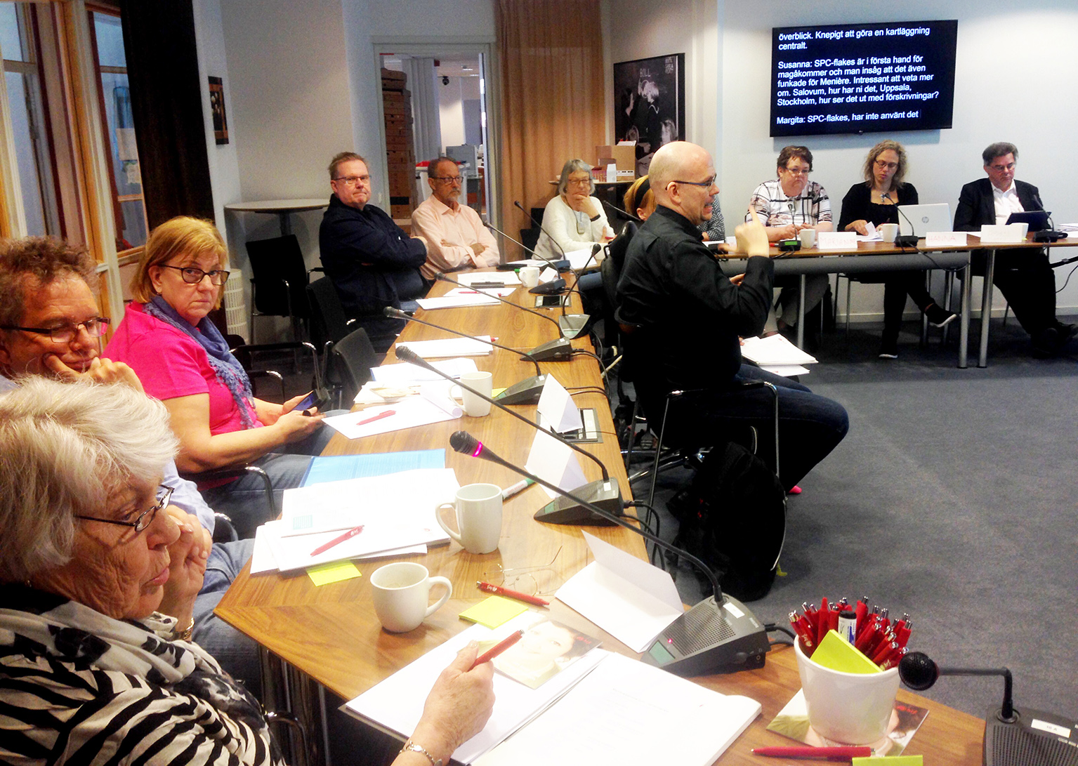 Personer sitter vid konferensbord med mikrfoner, med skrivtolkning på skärm