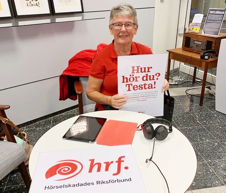 Kvinna i röd HRF-tröja håller upp skylt med texten "Hur hör du? Testa!"
