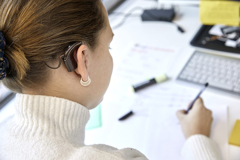 Kvinna med cochleaimplantat sedd snett bakifrån, framför skrivbord.
