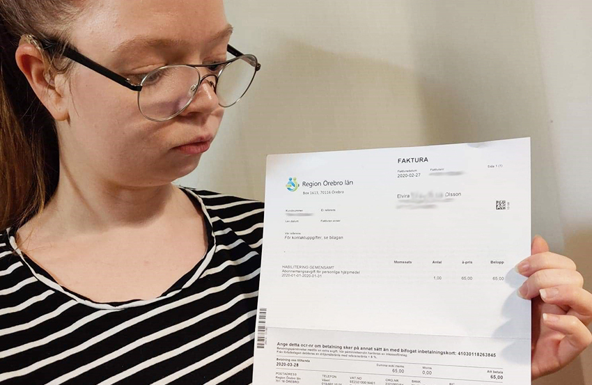 Ung kvinna med hörapparat och glasögon håller upp en faktura på 65 kronor från Region Örebro län för "abonnemangsavgift för personliga hjälpmedel".