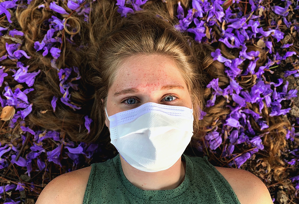 Kvinna med munskydd. Huvudet vilar på bädd med blå blommor.