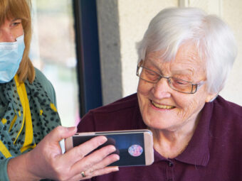 Äldre kvinna tittar på mobil som hålls fram av personal med munskydd.