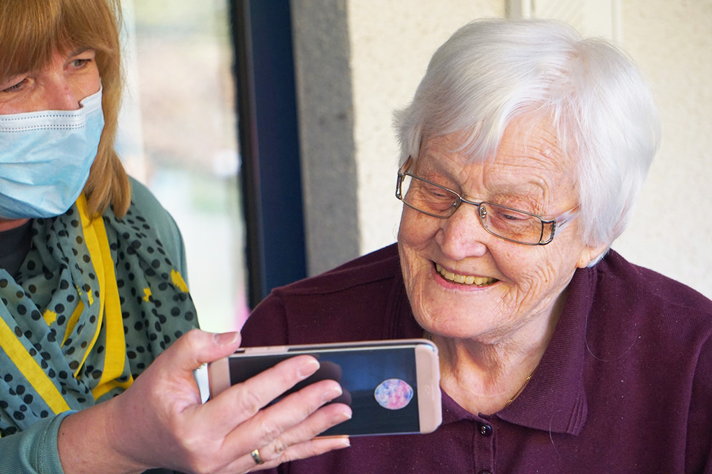 Äldre kvinna tittar på mobil som hålls fram av personal med munskydd.