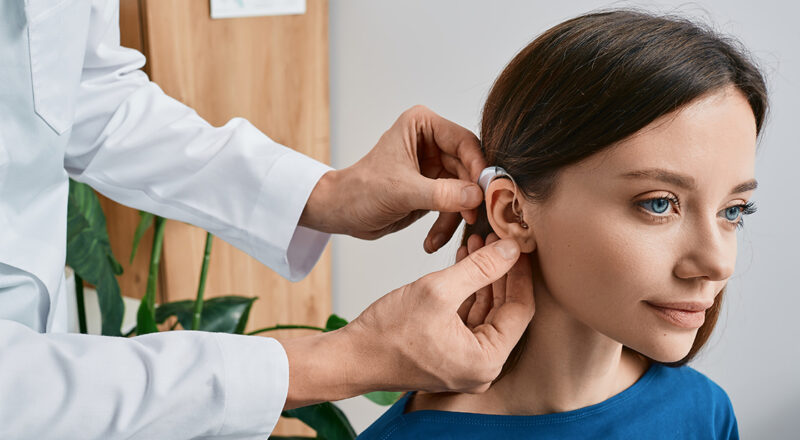 Kvinna med mörkt hår och blå tröja får en hörapparat placerad bakom örat av person med vit rock.