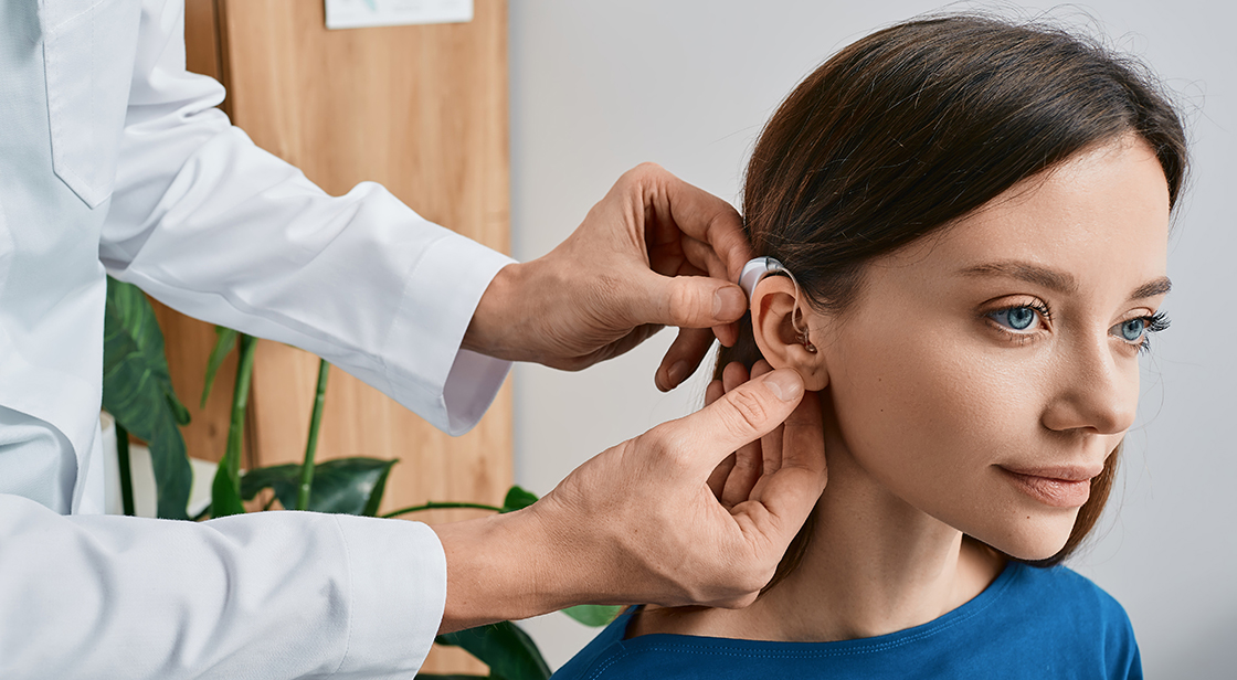 Kvinna med mörkt hår och blå tröja får en hörapparat placerad bakom örat av person med vit rock.