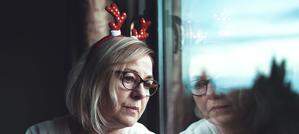 Kvinna i 60-årsåldern med rött renhornsdiadem på huvudet tittar ut genom fönster och ser tankfull och ledsen ut.