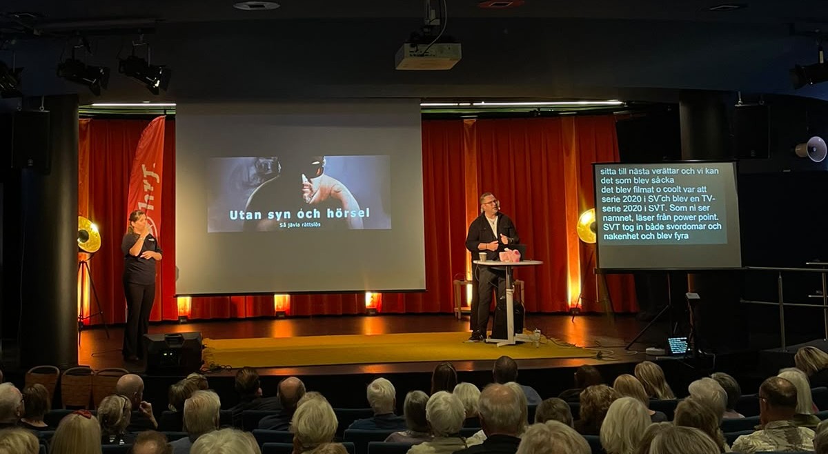 Torbjörn Svensson står på scen och berättar inför publiken. Teckenspråkstolk till vänter på scen och skrivtolkning syns på skärm till höger.